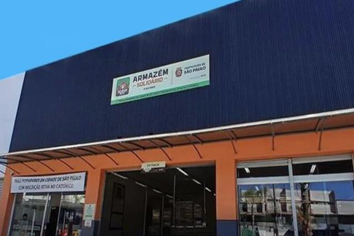  A imagem apresenta um estabelecimento dividido em duas partes. A parte de baixo laranja com uma entrada para o estabelecimento e com duas janelas localizadas cada uma em um lado. A parte superior tem a cor azul escura e contém um título ‘Armazém Solidário’, e ao lado direito da frase, o logo da prefeitura de São Paulo. 