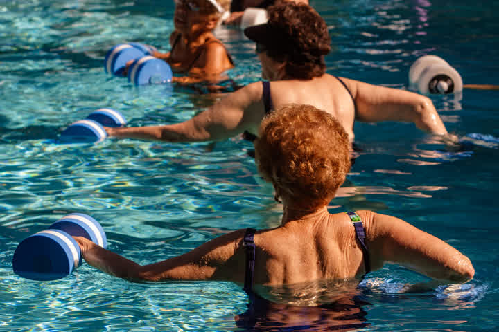 A imagem apresenta um grupo de pessoas idosas se exercitando com pesos em uma piscina.