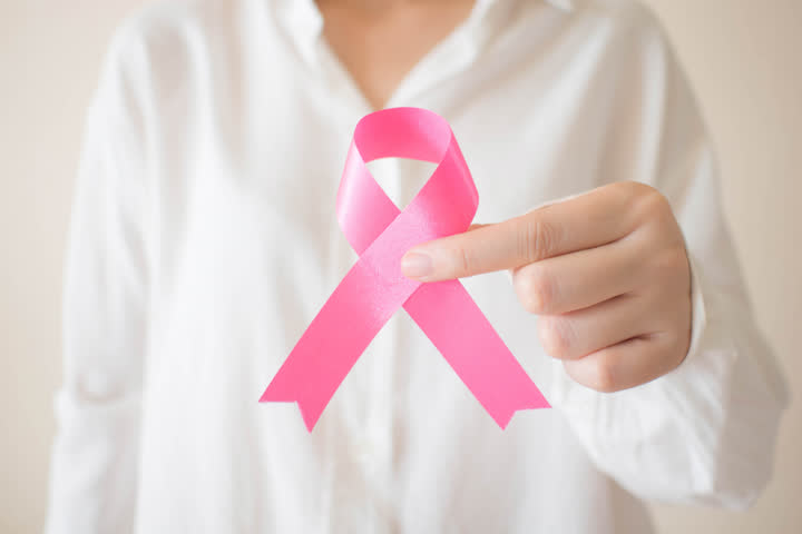 Na imagem pode-se ver a parte do tronco de uma mulher de cor branca, vestida com uma camisa de manga longa branca. A frente do seu corpo ela segura um laço na cor rosa.
