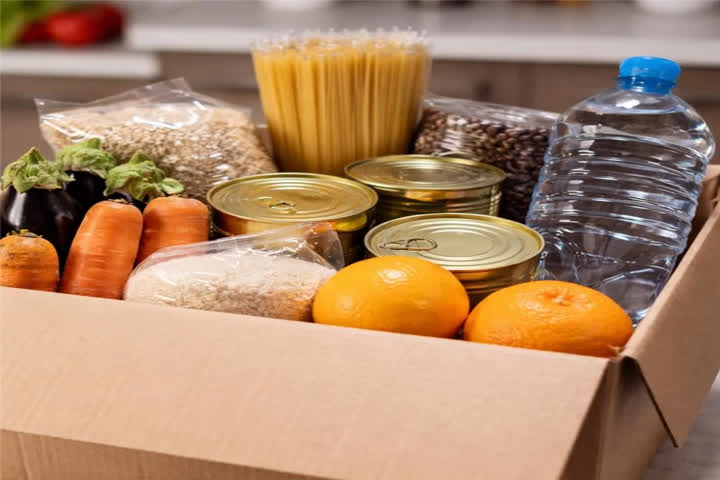 A imagem de uma cesta básica organizada dentro de uma caixa de papelão aberta, apoiada sobre uma bancada de cozinha clara. Dentro da caixa há alimentos variados.