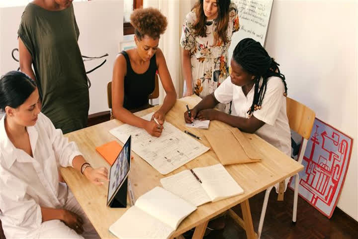 Na imagem há uma mesa retangular de madeira clara, ao redor da qual cinco pessoas participam de uma atividade colaborativa.