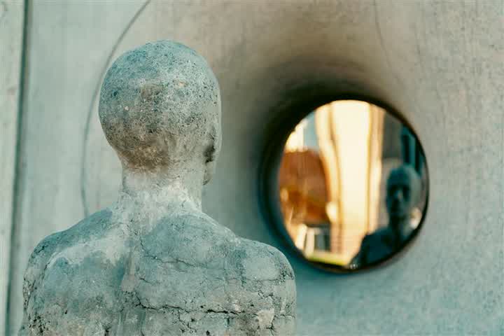Imagem em ambiente externo com foco em uma escultura humana vista por trás. O reflexo está levemente desfocado, e ao fundo é possível perceber formas e cores suaves que sugerem construções ou elementos urbanos.