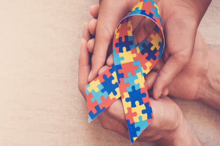 Imagem em plano aproximado de duas mãos adultas segurando delicadamente as mãos de uma criança. As mãos estão posicionadas no centro da cena, transmitindo sensação de cuidado e acolhimento. Entre elas, há um laço em formato de fita, com estampa colorida de peças de quebra-cabeça nas cores azul, vermelho, amarelo e verde.