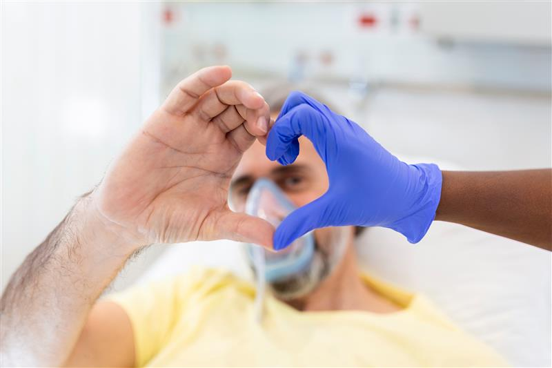 A imagem apresenta uma pessoa de cor branca, do sexo masculino, em ambiente hospitalar, com a mão direita a frente do corpo formando um coração, junto com a mão de uma pessoa de cor preta, vestida com uma luva de procedimento na cor azul.