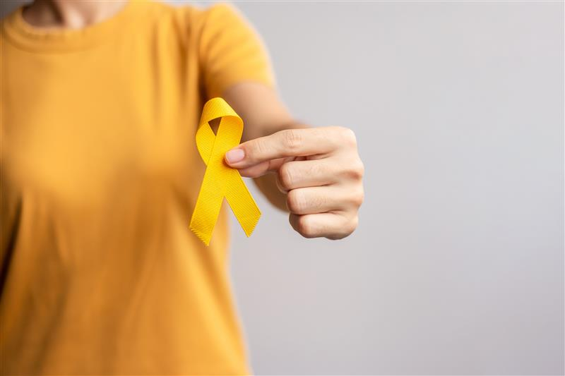 A imagem apresenta o tórax de uma pessoa de cor branca, vestida com uma blusa de cor amarela, com a mão esquerda a frente do corpo, segurando um laço amarelo.